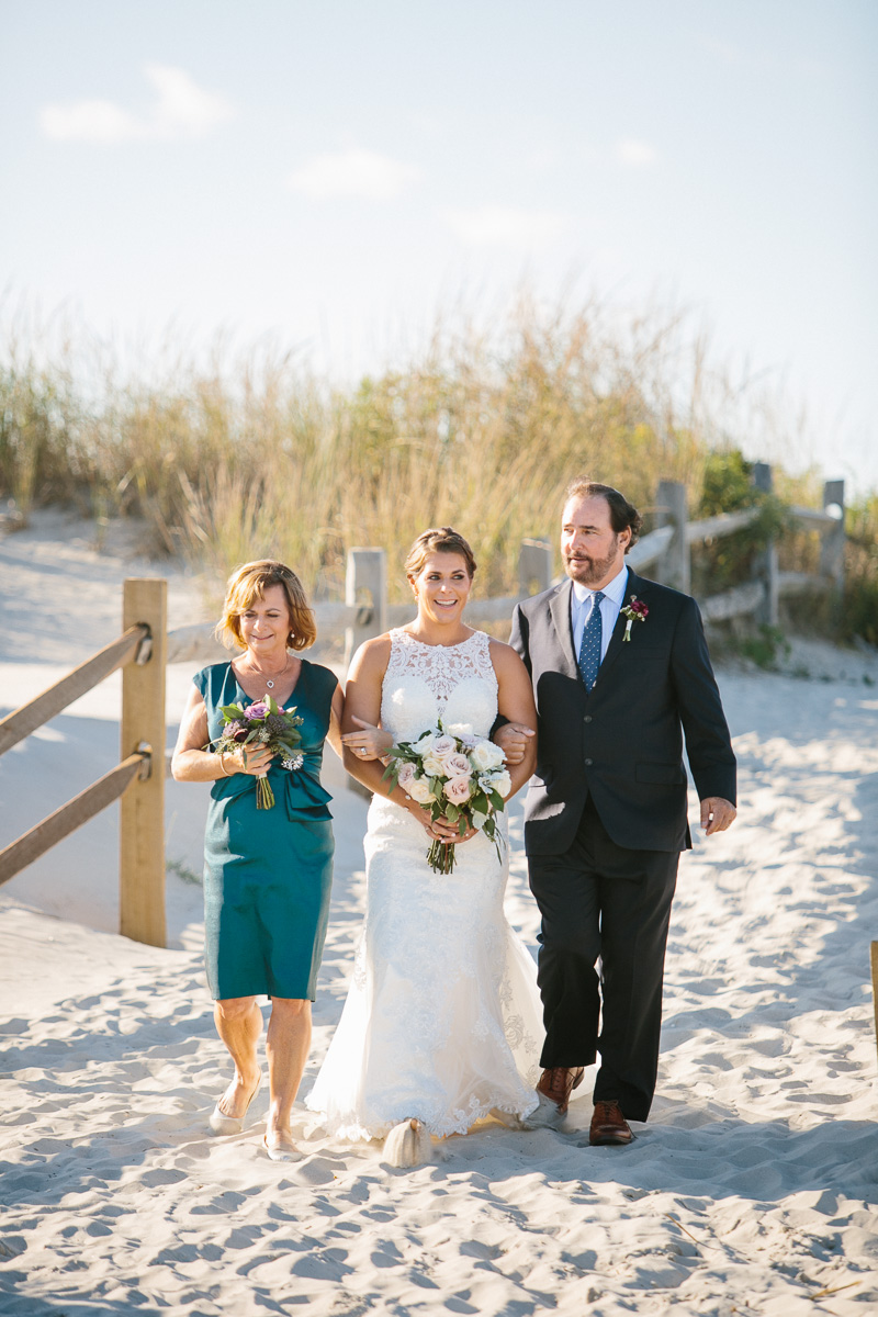Bride walks down the aisle with parents during outdoor, beach wedding ceremony.