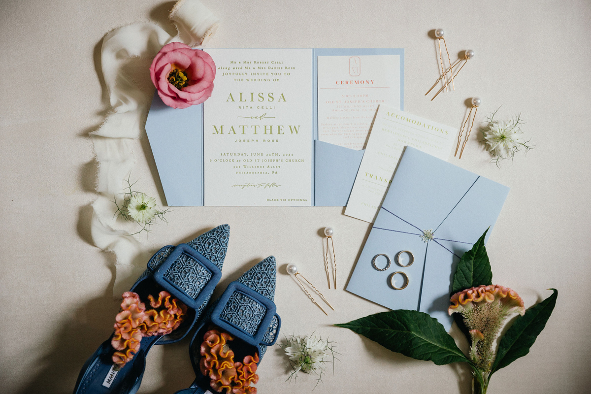 Flatlay of wedding invitation suite with blue heels, and pink and orange florals.