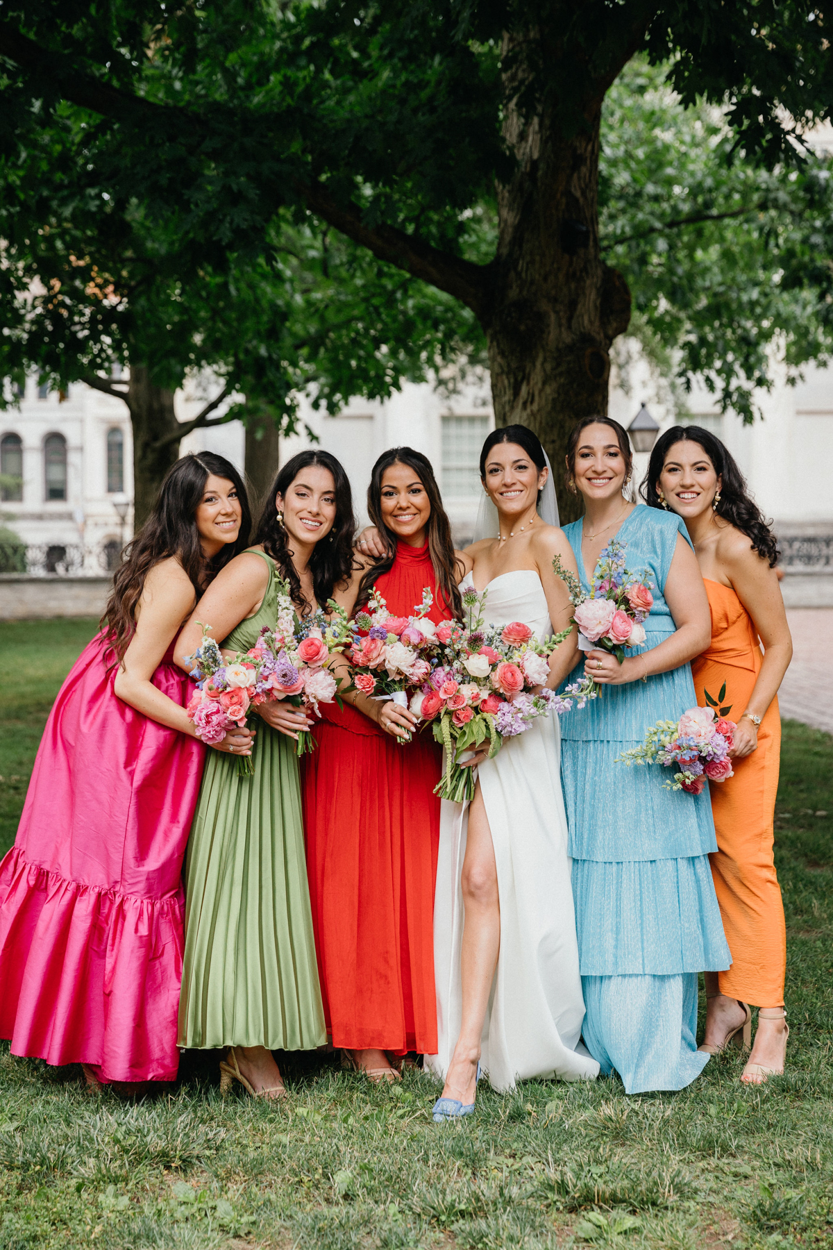 Bridesmaids in mismatching dresses. From left to right the dress colors are, hot pink, olive green, red, white, light blue, and orange.