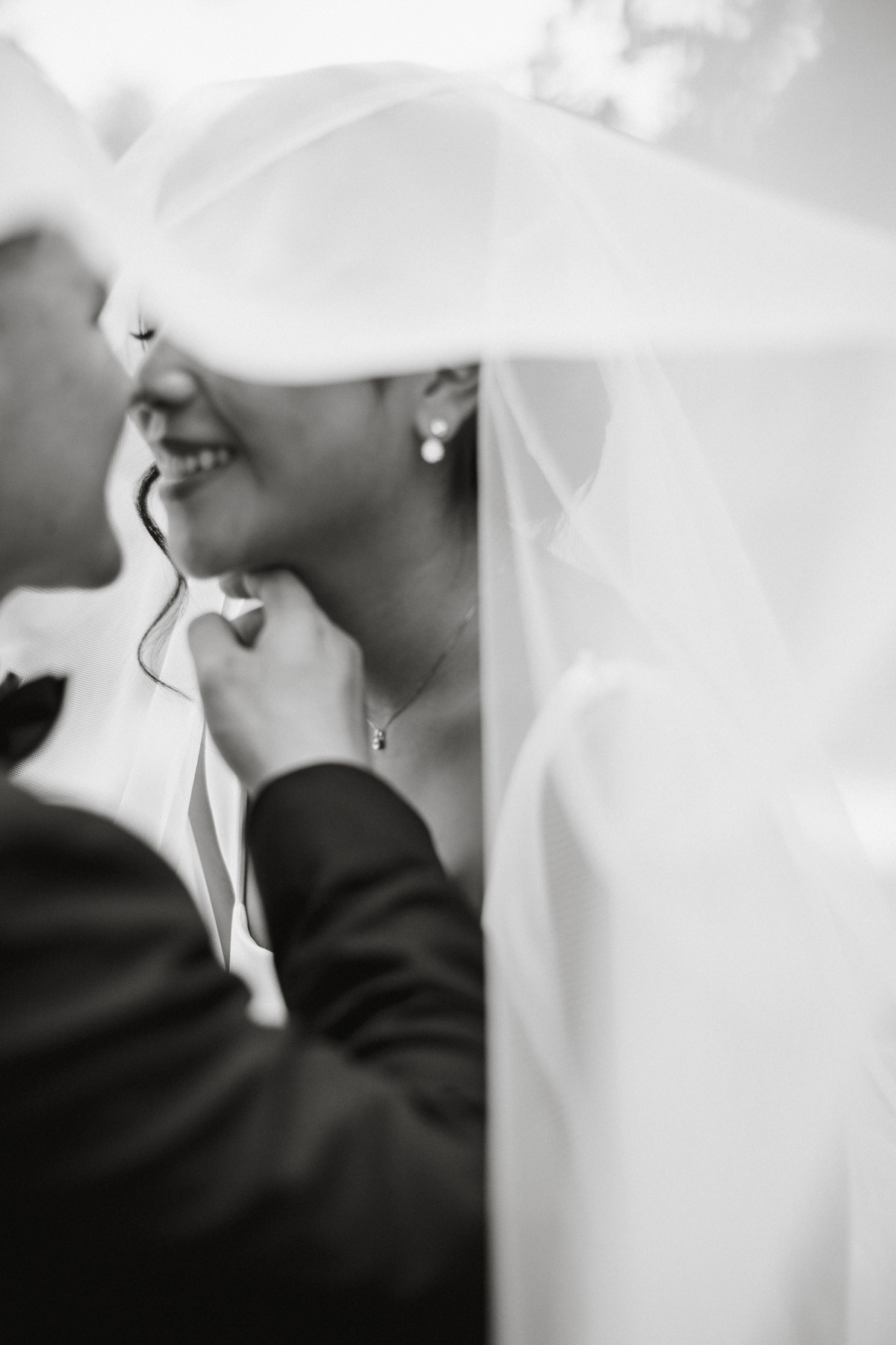 Black and white photo of bride and groom smiling, about to kiss, underneath her veil.