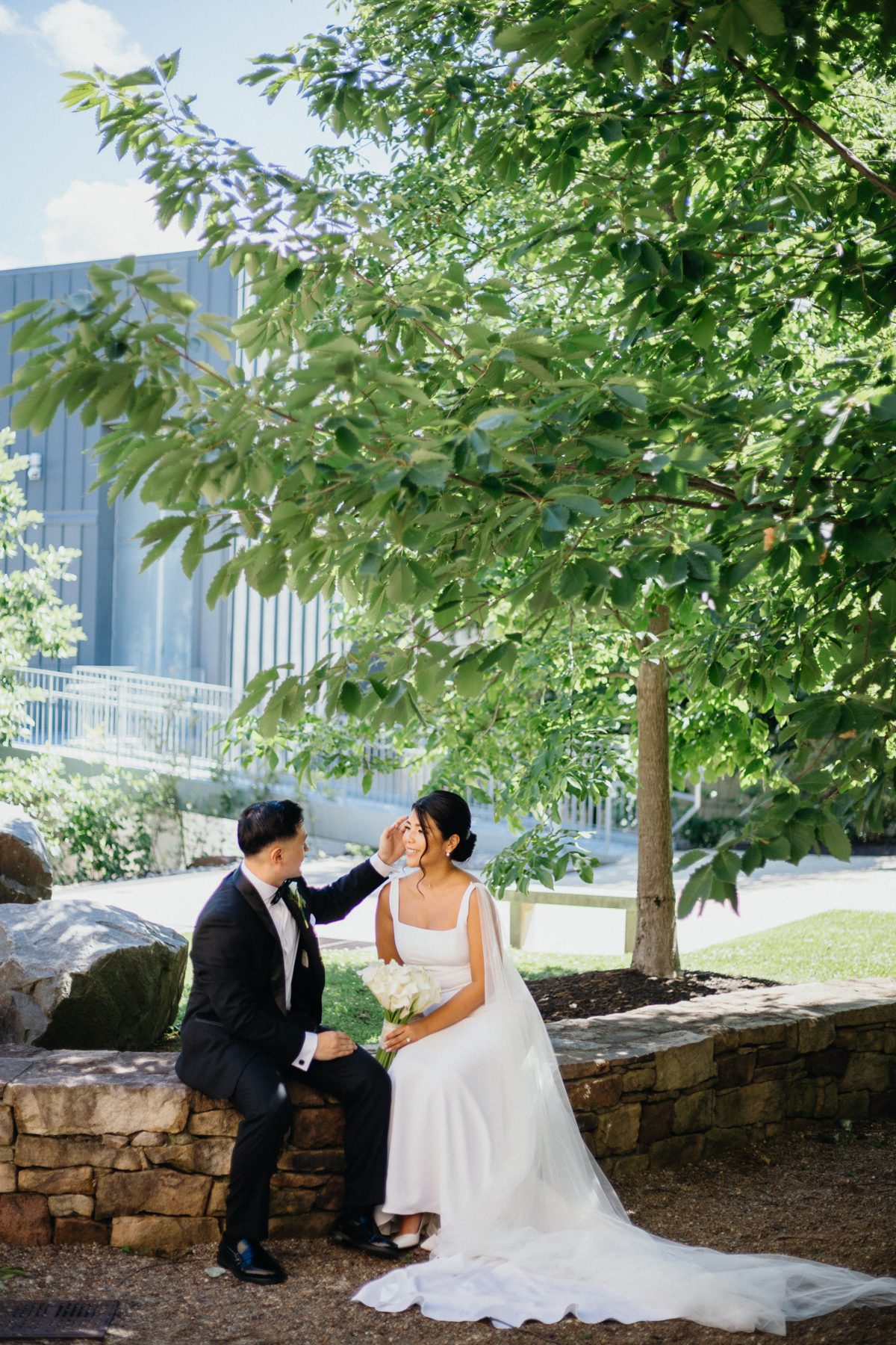 Groom brushing hair away from brides face as they sit on a stone wall outside a museum.