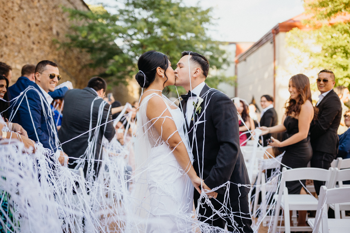 Bride and groom kiss covered in white streamers as they exit outdoor sculpture garden wedding ceremony.