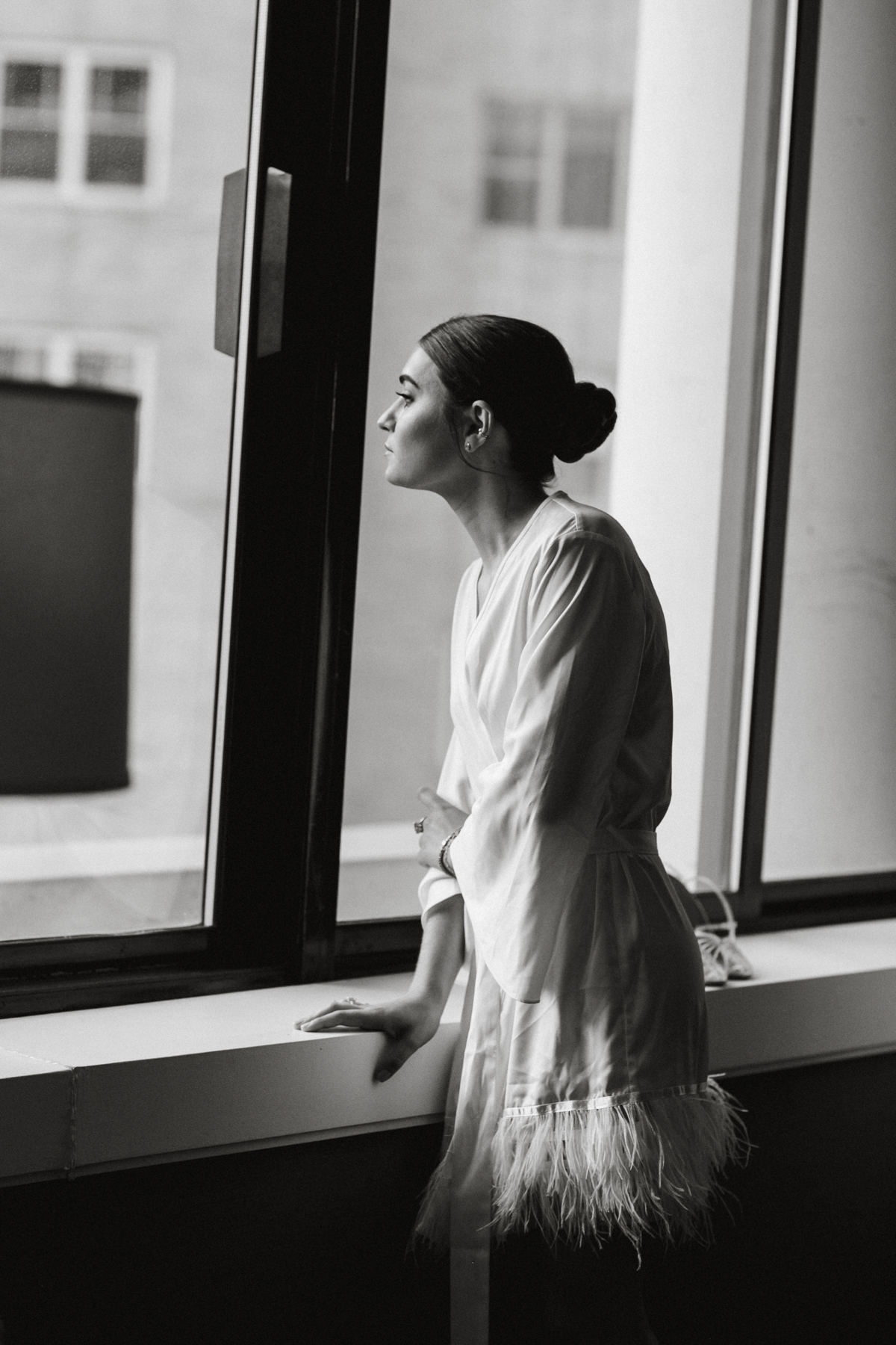 Image of bride in a robe with a slick back bun staring out a window.