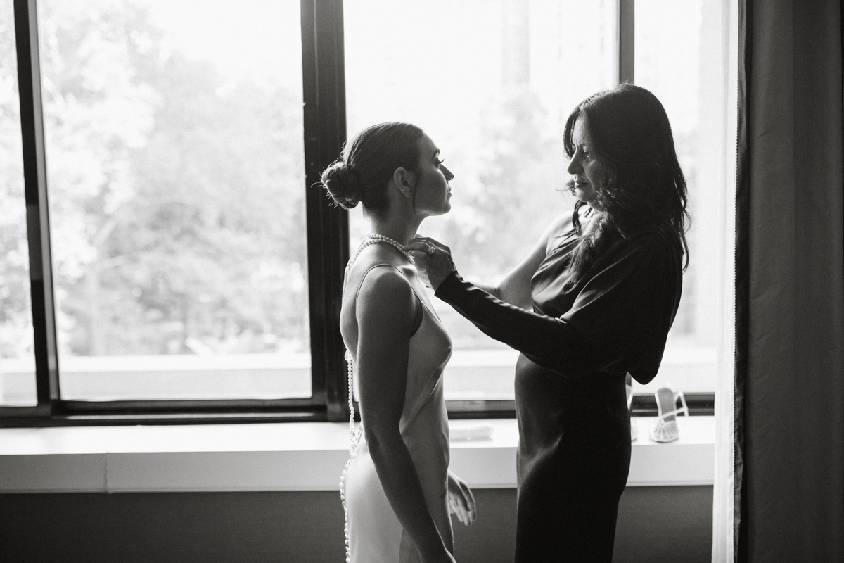 Black and white image of mom assisting bride with necklace on the morning of her wedding.