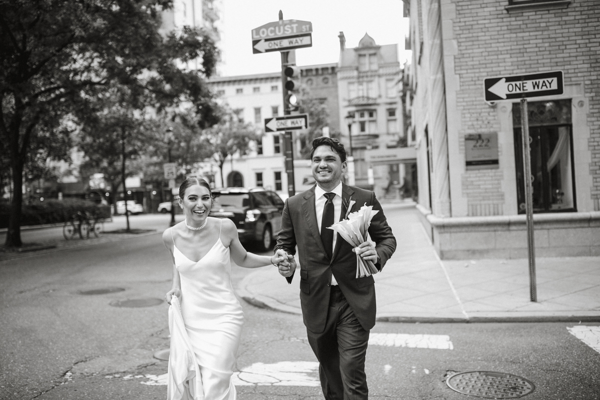 Black and white of a wedding couple laughing and walking across a crosswalk.