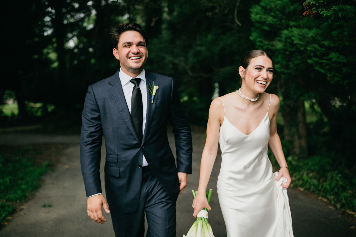 Bride and groom laughing as they walk through trees and greenery.