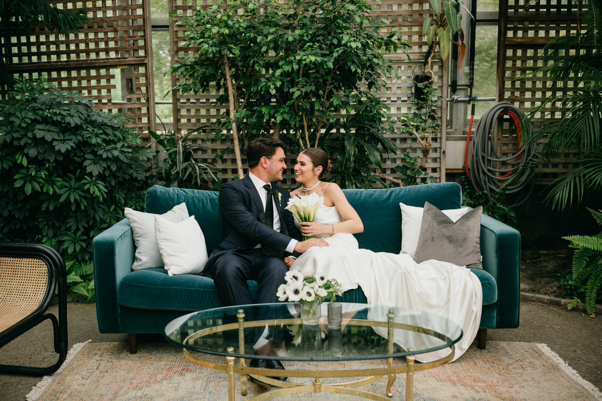 Wedding couple sitting on emerald couch smiling at each other.