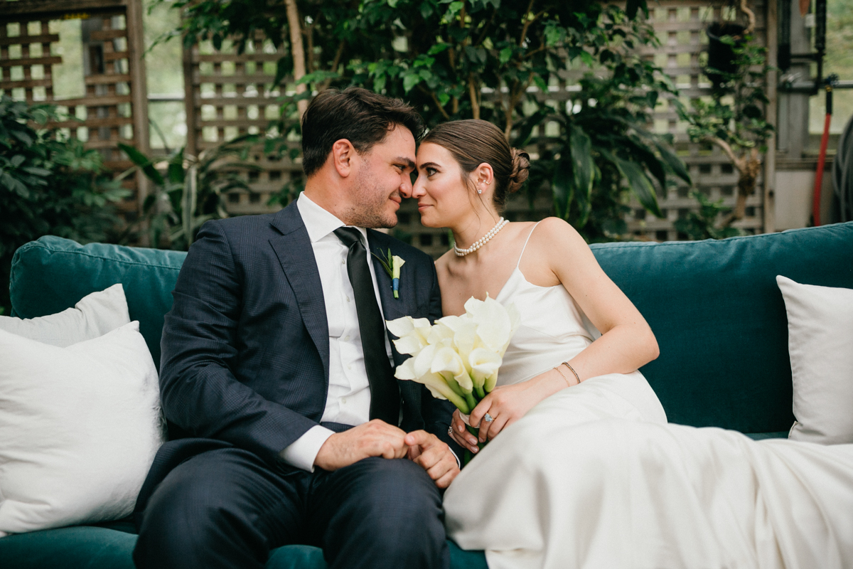 Close up of couple sitting on a green couch, smiling at each other on their wedding day.