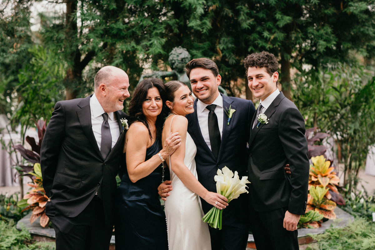 Bride and groom laughing with family in a green house wedding venue.