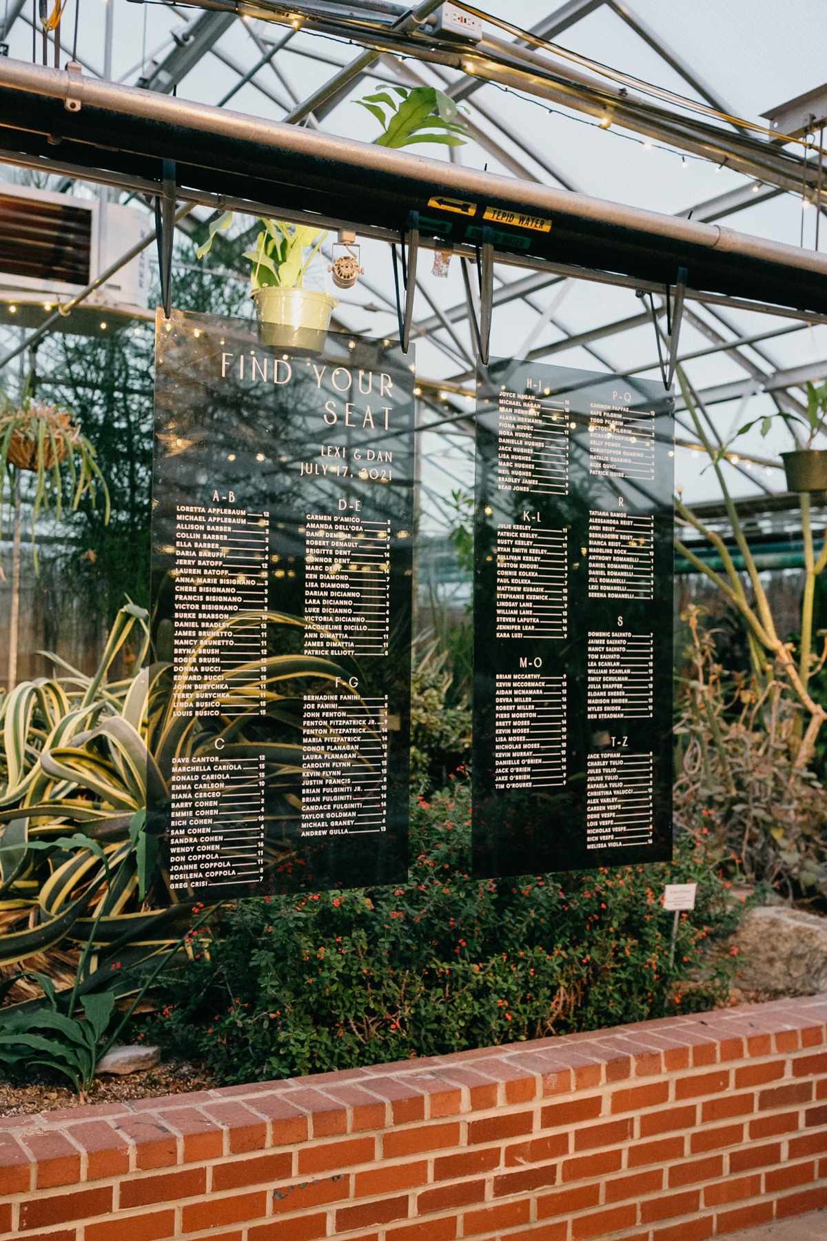 Clear, glass wedding seating chart hung in front of lush greenery.