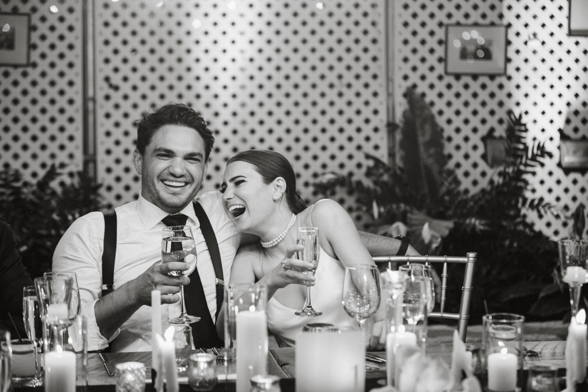 Black and white of wedding couple laughing as they sit at their table for dinner.