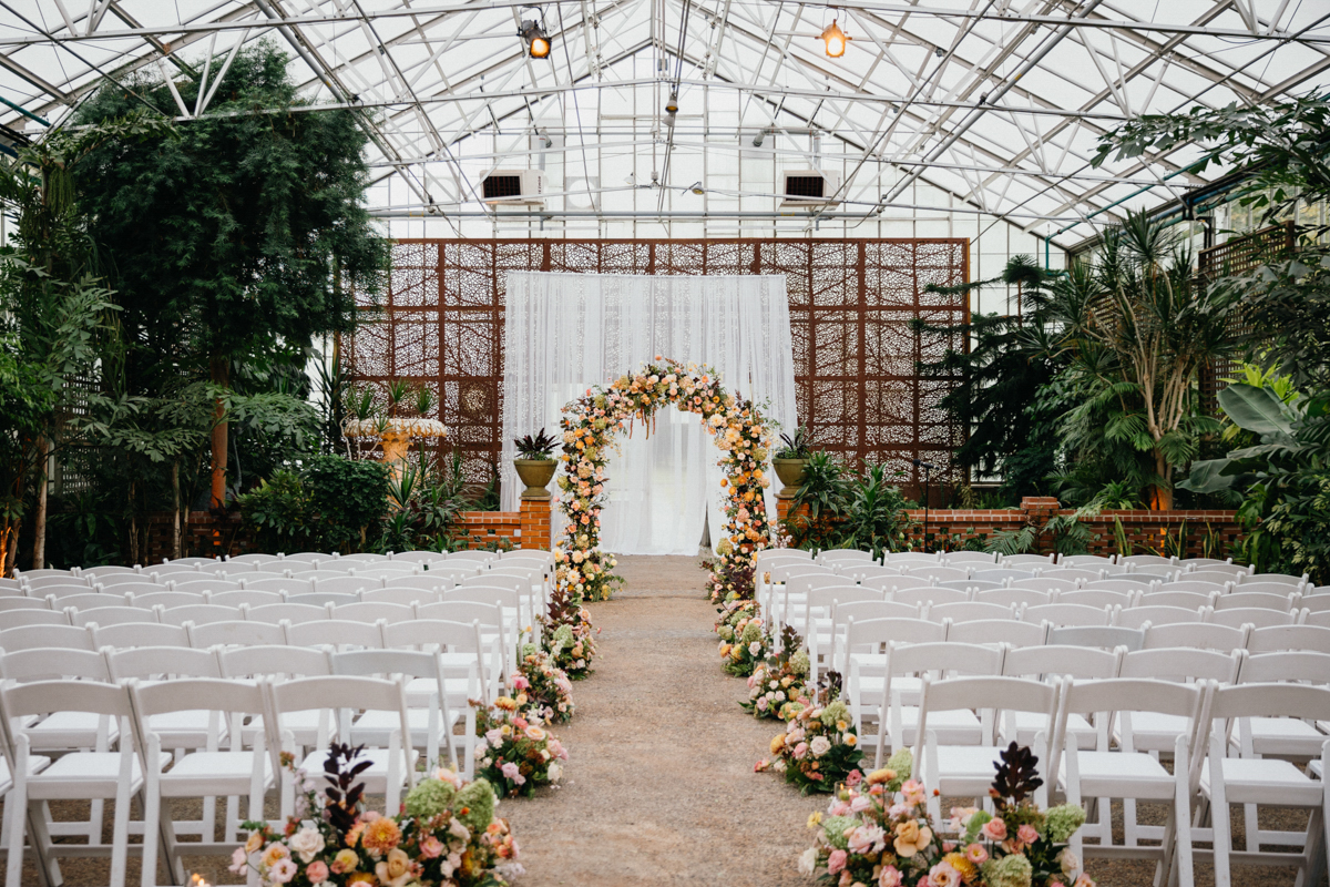 Vibrant summer wedding florals up the aisle and in an arch at the altar in a greenhouse wedding venue.