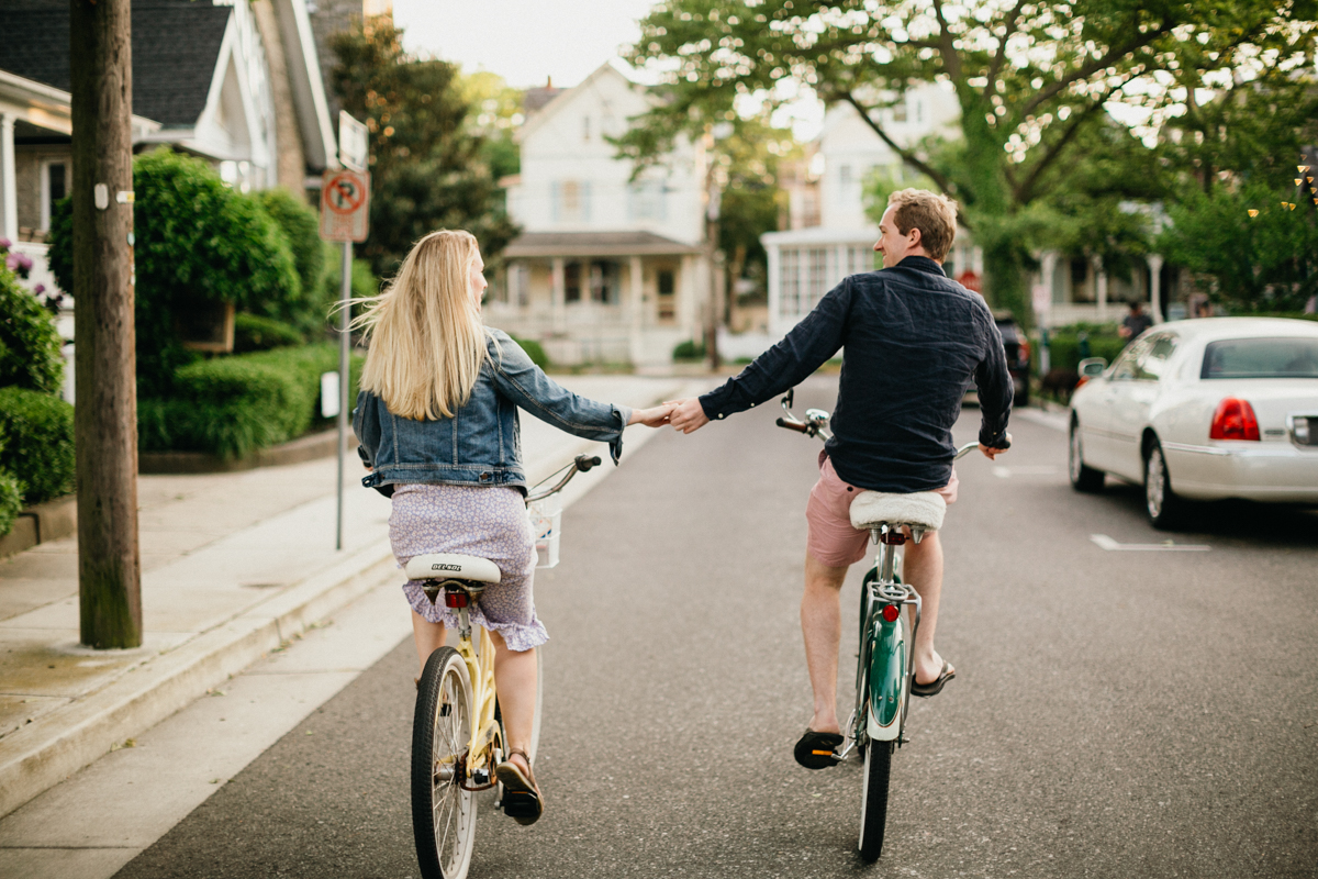 A couple pedals down a charming street in Cape May, surrounded by Victorian homes and lush greenery.