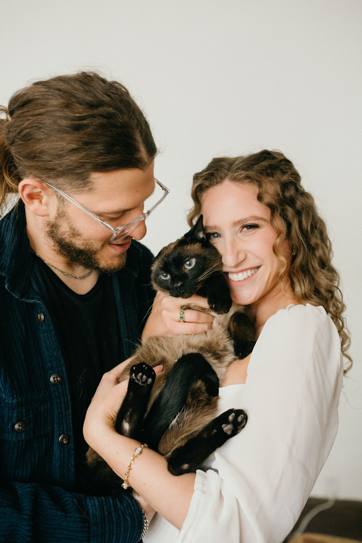 A couple smiles as they hold their cat, lounging in bed and sharing a peaceful moment together.