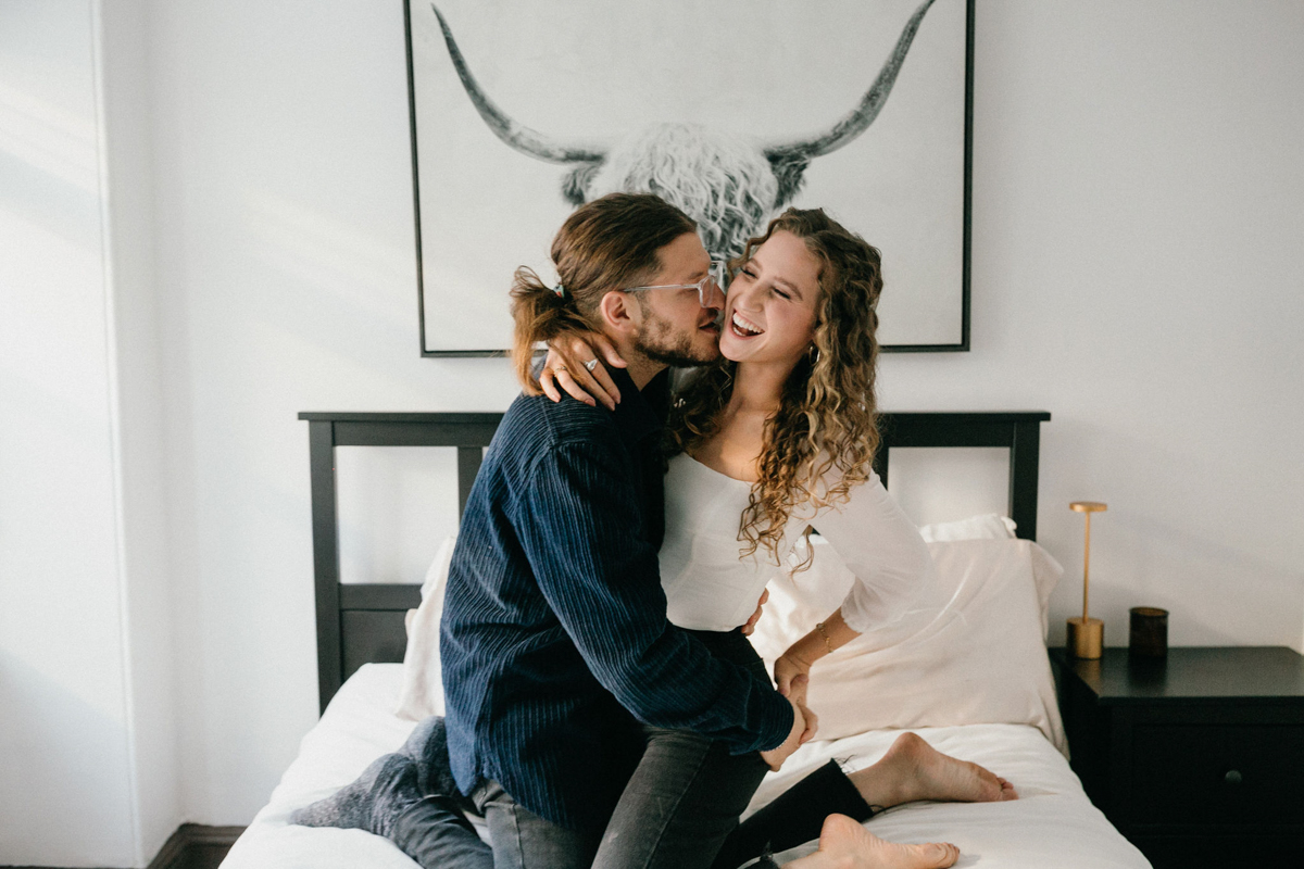 Couple laughs and plays in bed during an at home engagement session in Philadelphia, PA. .