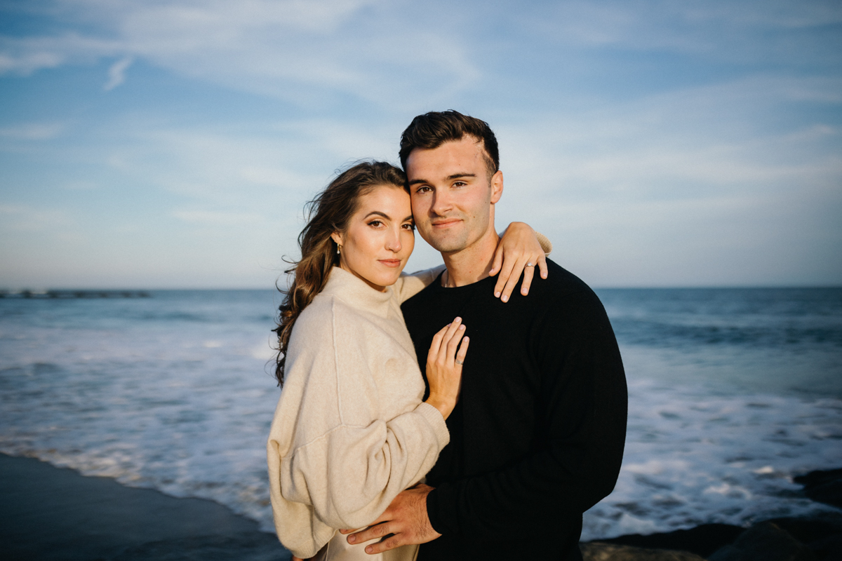 A couple stands on a sandy beach, with the waves of the Jersey Shore in the background, sharing a quiet moment.