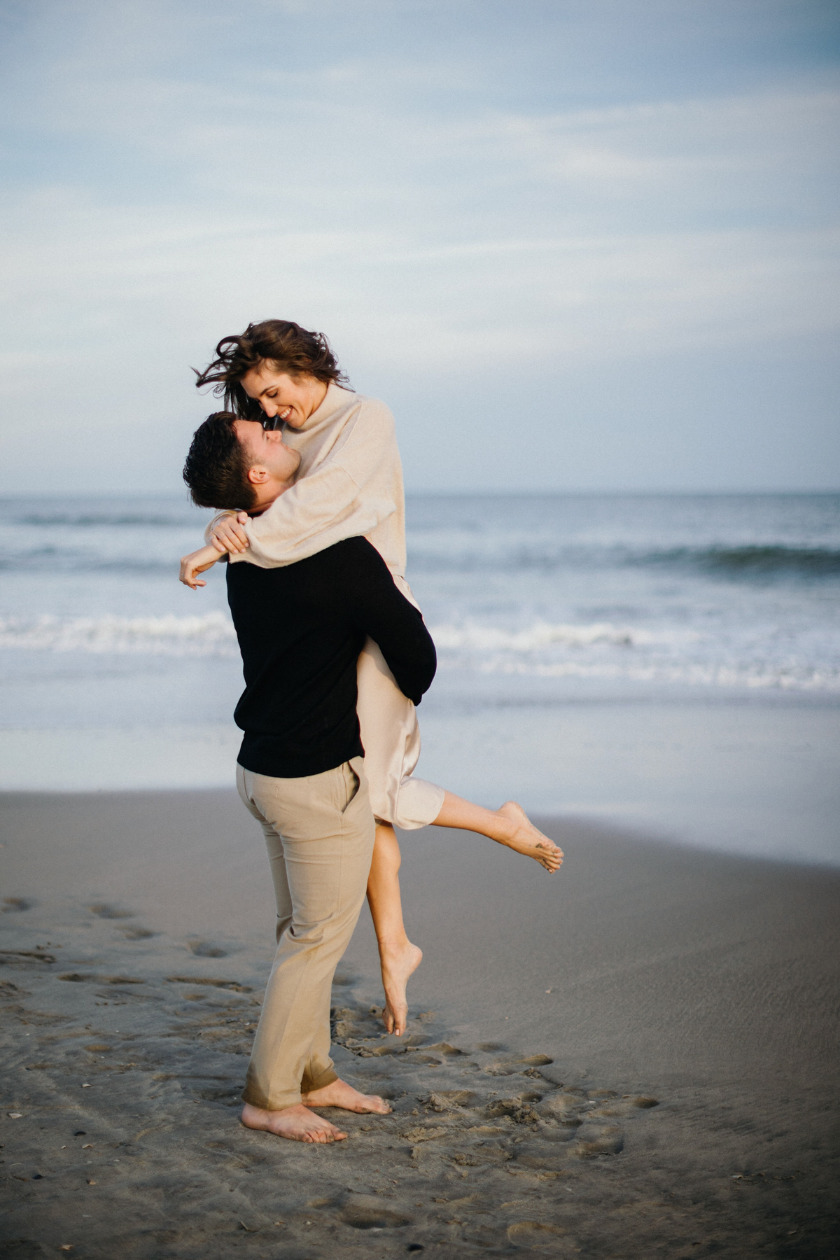 A couple poses barefoot near the shoreline, with the ocean waves gently crashing behind them.