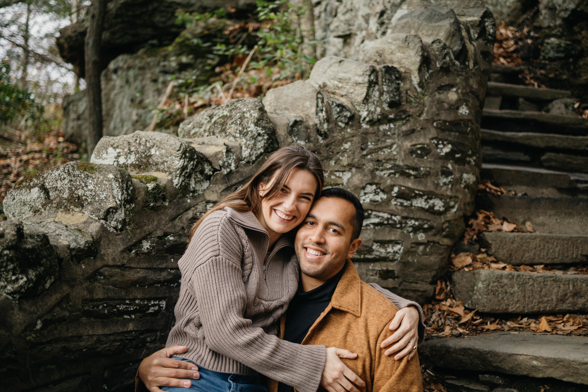 A couple poses near a towering tree with textured bark in a forest near Philadelphia, surrounded by fallen leaves.