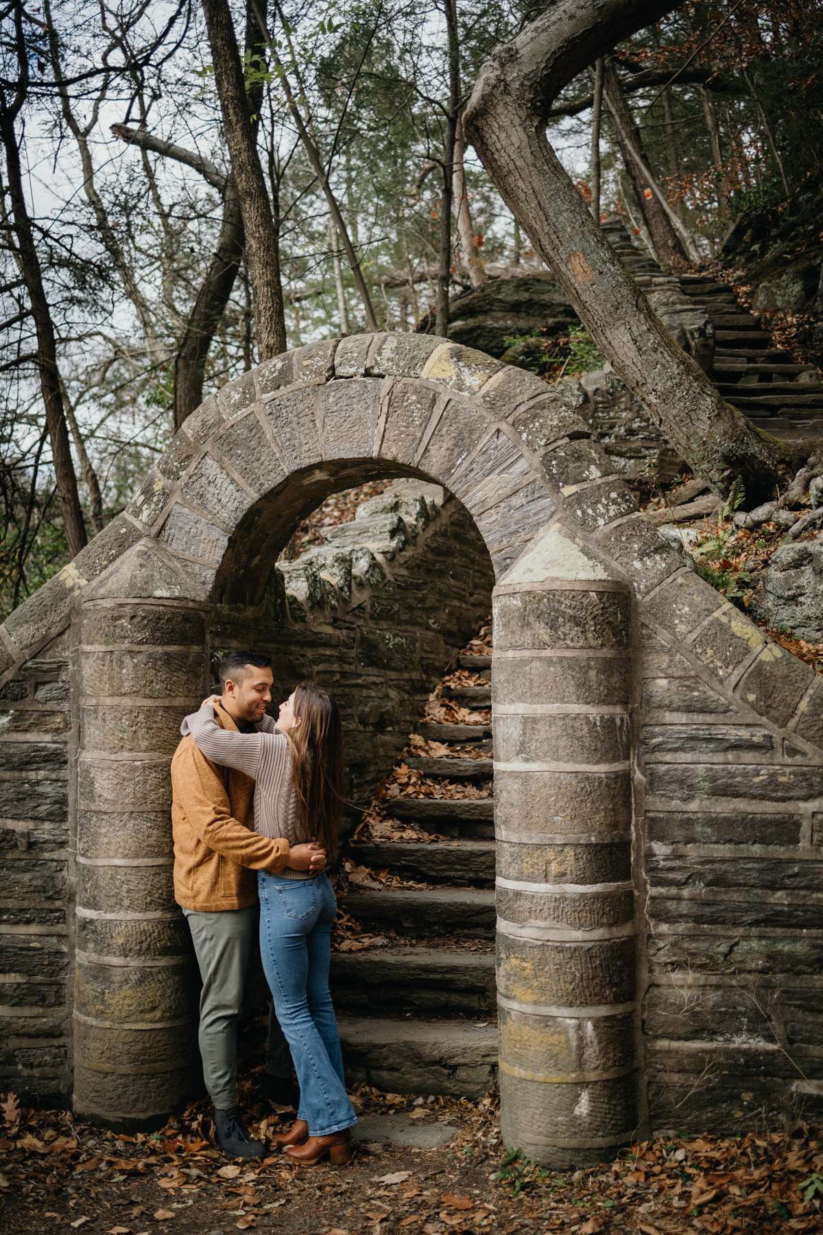 A couple sits on a mossy log, framed by dense greenery and soft light filtering through the trees near Philadelphia.