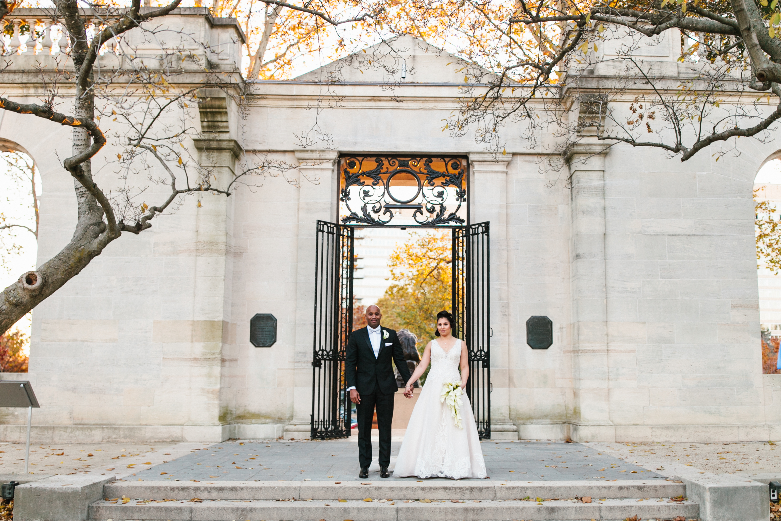 Newlyweds walk hand in hand through a grand courtyard, surrounded by sculptures and lush greenery.