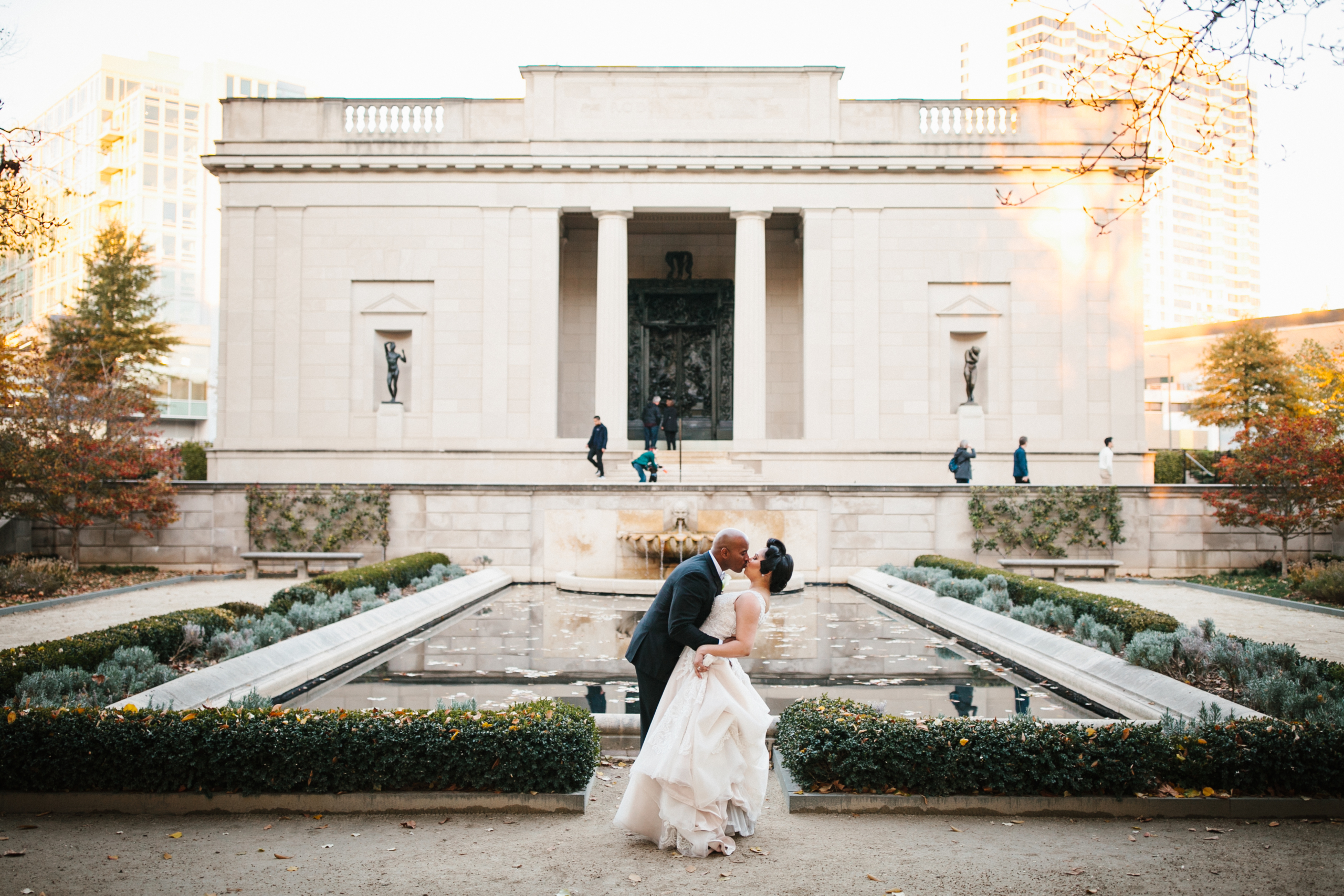 The couple shares a quiet moment near a stately stone fountain, soft light filtering through the trees.