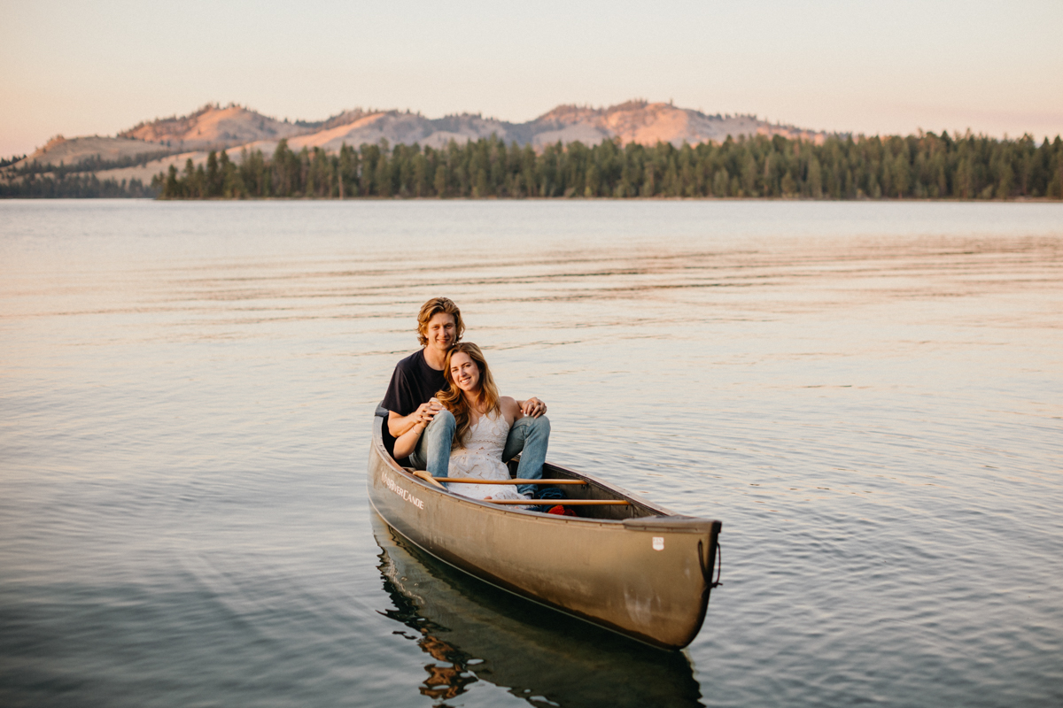 A couple paddles together in a bright kayak on a calm lake surrounded by trees.