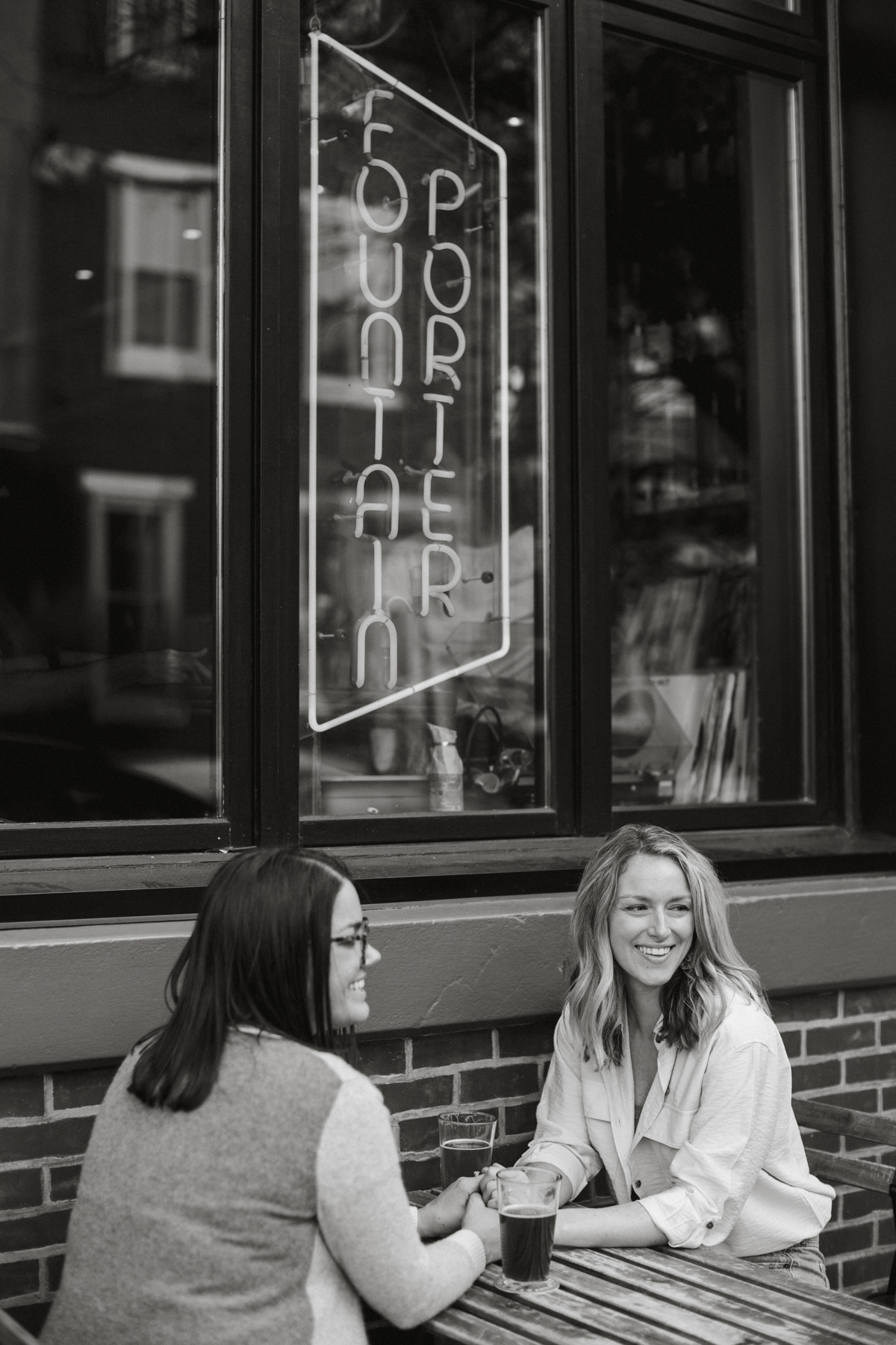 A couple poses near the neon sign of a trendy bar in South Philly, with drinks in hand and soft evening light.