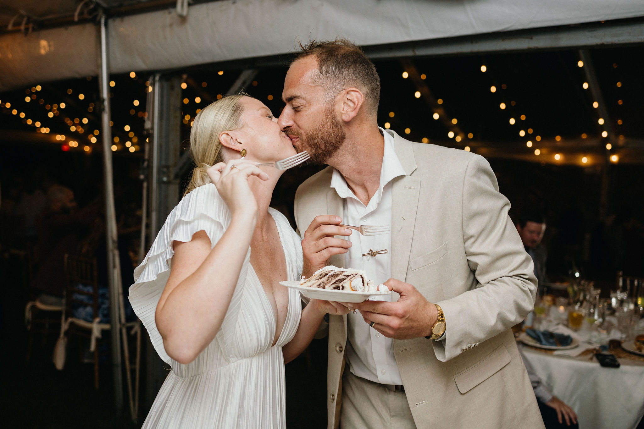 String lights glowing over the outdoor wedding reception as the bride and groom share a kiss.