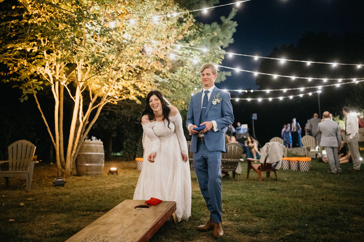 A bride and groom enjoying a round of cornhole under string lights.