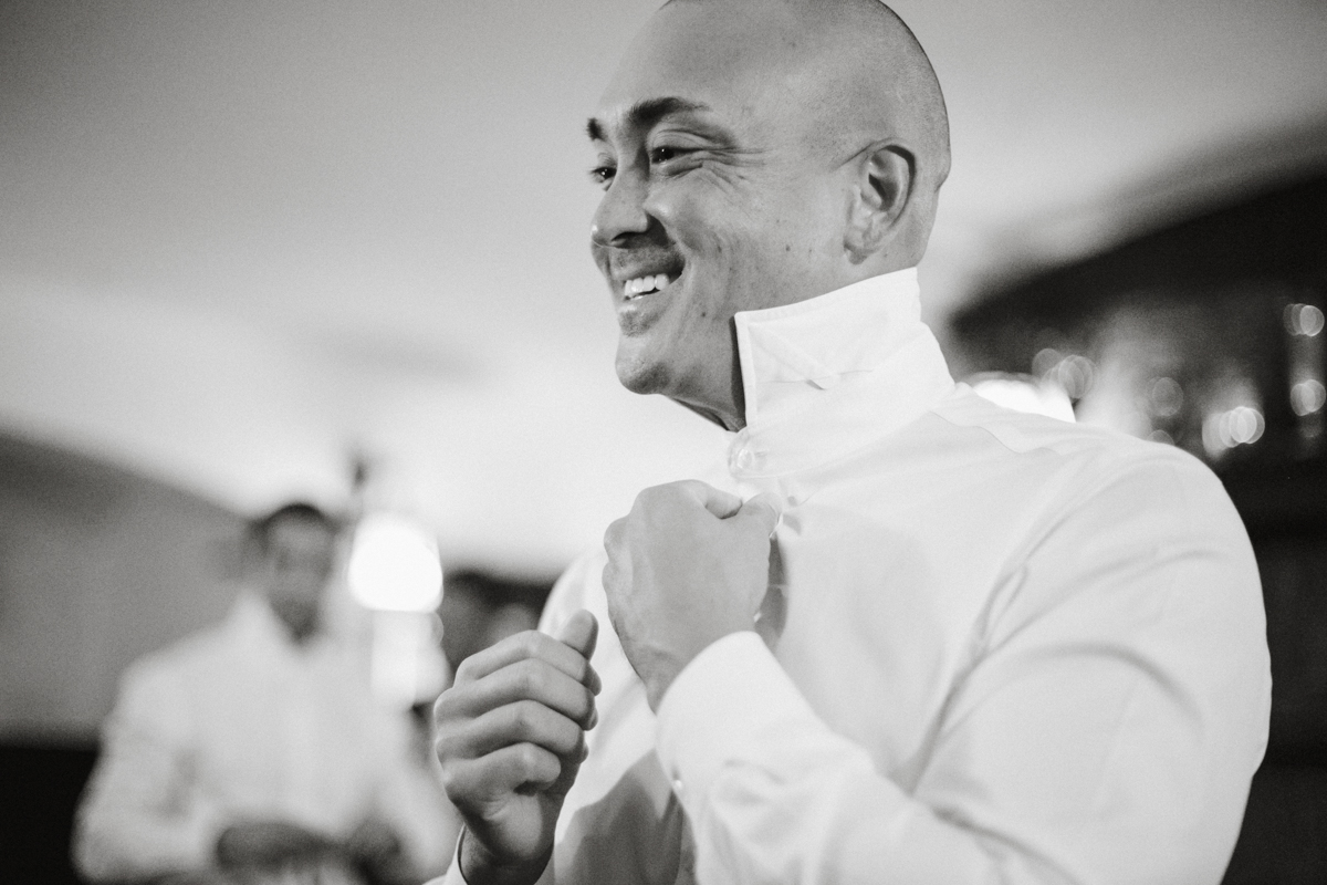 Groom adjusting tie by riverfront window – editorial wedding photography