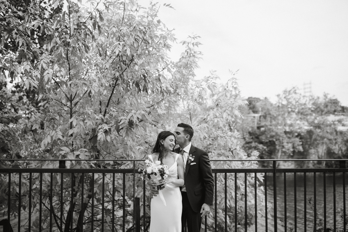 Groom kissing bride’s forehead on waterfront at Pencoyd